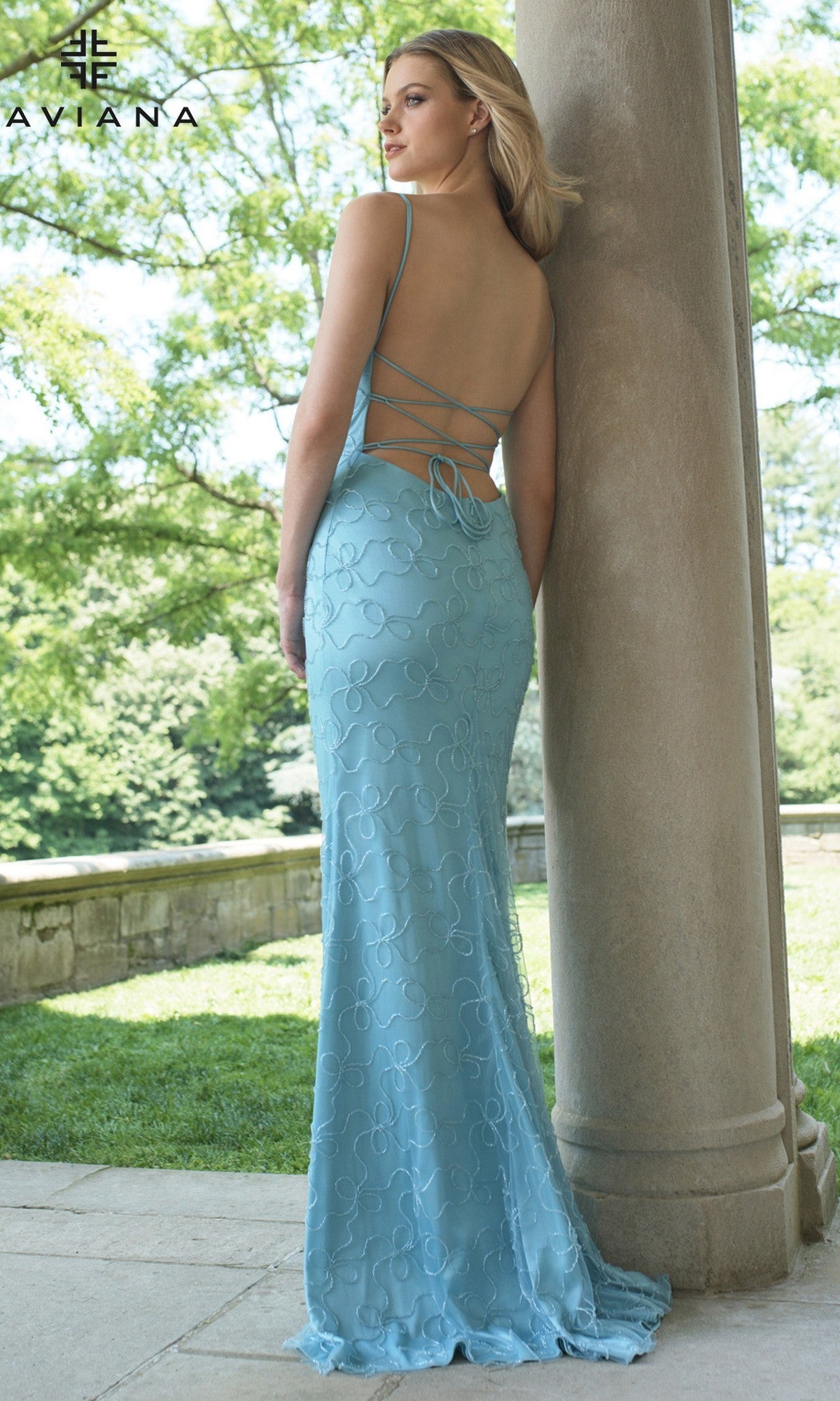 A woman stands outdoors next to a stone column wearing the Faviana 11417 long prom dress in light blue, featuring a v-neckline, thin straps, and a lace-up back. Trees and a stone wall are visible in the background.
